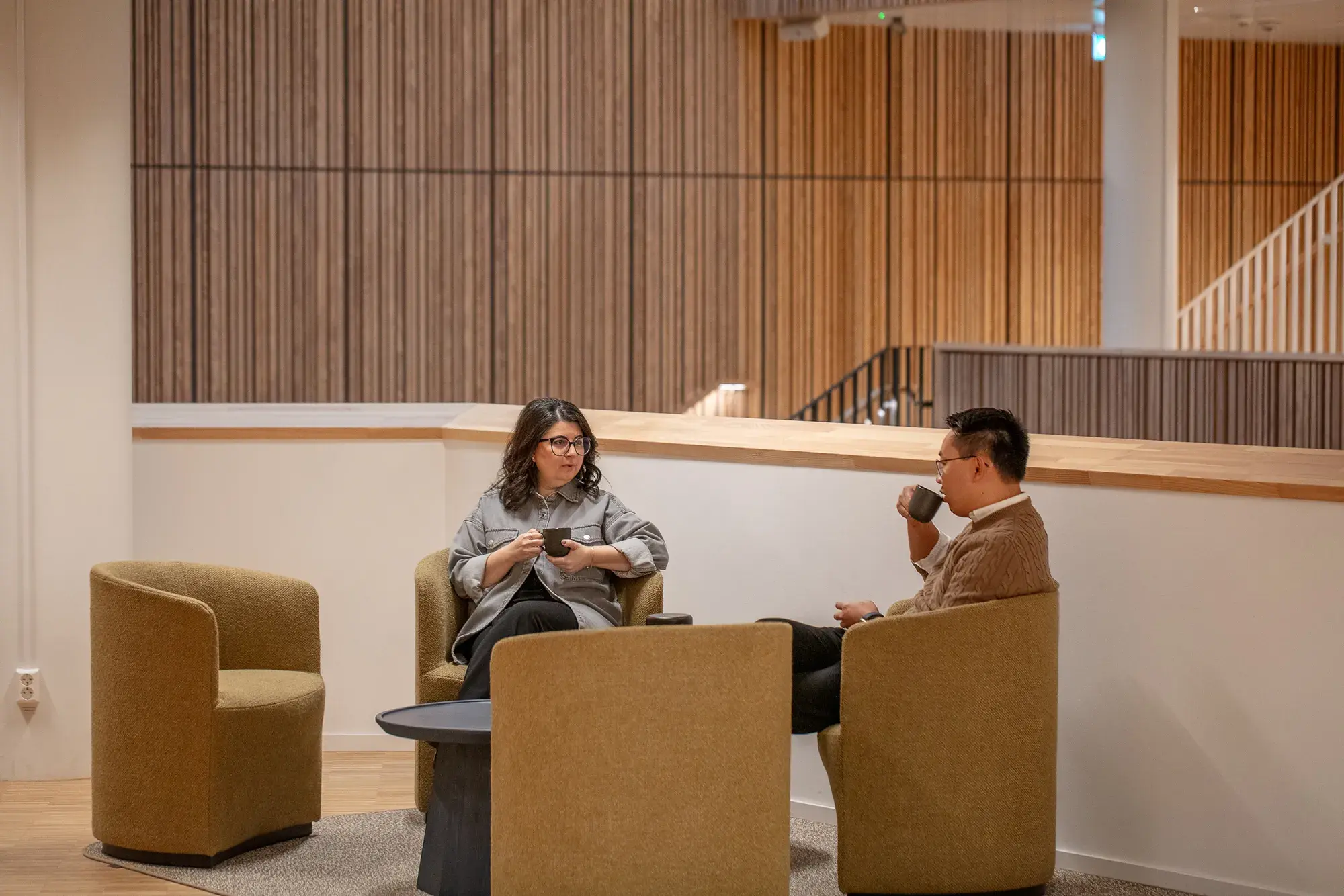 Two people sitting in beige chairs, drinking coffee and conversing in a modern setting with wood paneling.