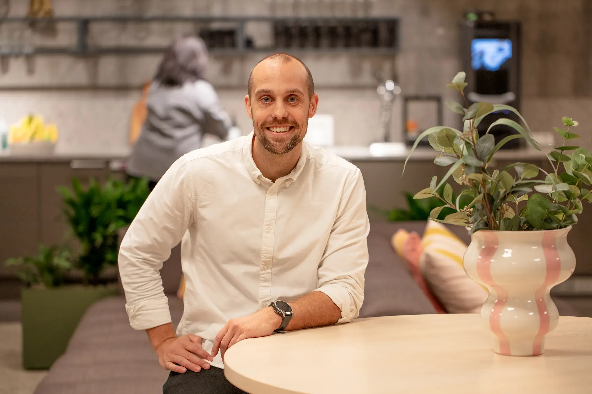 Smiling man in a white shirt sitting at a table with a pink-striped vase, plants, and a woman in the background in a kitchen setting.