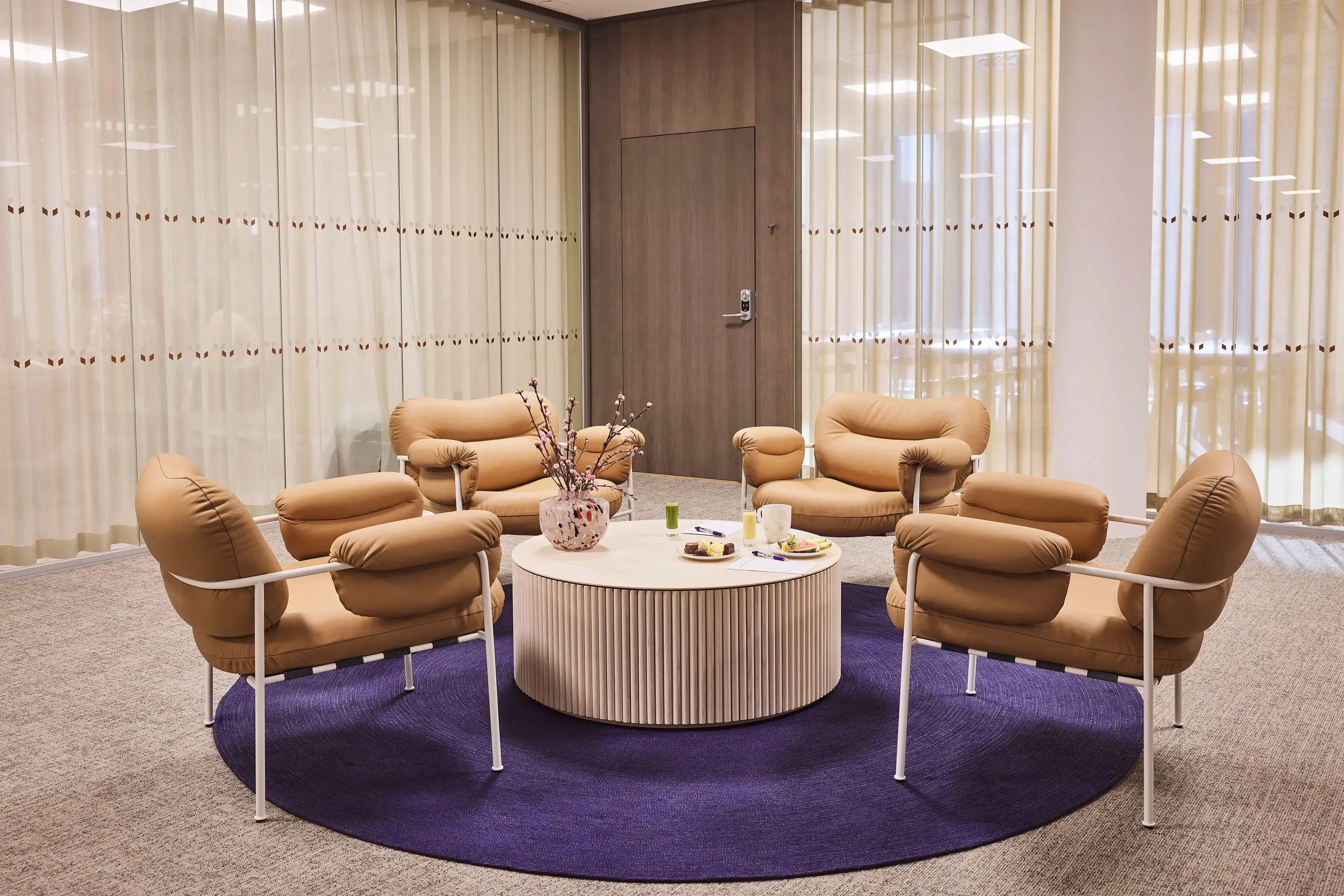 Cozy seating area with tan armchairs, round coffee table, and vase on blue rug, surrounded by beige curtains and wood panel door.